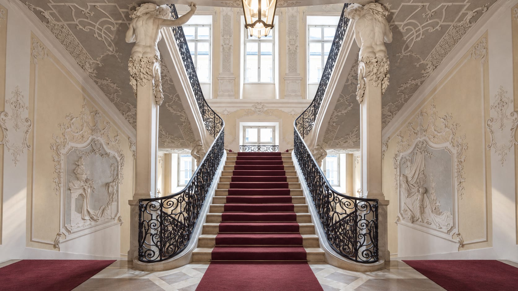 Treppenhaus im Palais Preysing: zweiläufige Treppe mit rotem Läufer, schmiedeeisernen Geländern und Stuckfiguren. Mit KI bearbeitete Darstellung.